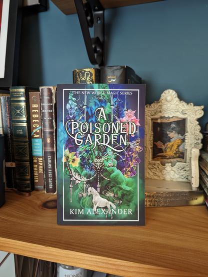 picture of the book A Poisoned Garden standing on a bookshelf. Behind that book, there are other books and an aged white picture frame with a picture of a woman in a yellow Victorian dress reading a book. Book cover of A Poisoned Garden is a colorful cover filled with green, pink, purple, blue flowers and smoke and a white horse on the bottom of the cover.
