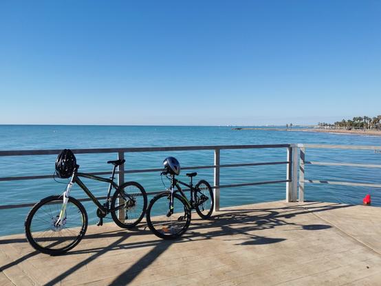 Se ven en primer plano dos bicicletas de montaña con un casco en cada manillar. Están apoyadas en una barandilla y al fondo se ve el mar y a la derecha de la toma se ve una playa al fondo.