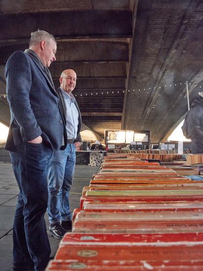 Das Bild zeigt eine Szene vom South Bank Used Book Market in London. Im Vordergrund sieht man lange Reihen von gebrauchten Büchern mit roten Einbänden, die auf Tischen ausgelegt sind. Zwei Männer mittleren Alters stehen daneben und betrachten die Bücher. Der vordere Mann trägt einen dunkelblauen Blazer und Jeans, der hintere ein hellblaues Hemd. Sie befinden sich in einer überdachten Umgebung, vermutlich unter einer Brücke, erkennbar an der gewölbten Decke über ihnen. An der Decke hängen Lichterketten, die eine gemütliche Atmosphäre schaffen. Im Hintergrund sind weitere Bücherstände und Besucher zu sehen, was auf einen belebten Markt hindeutet.