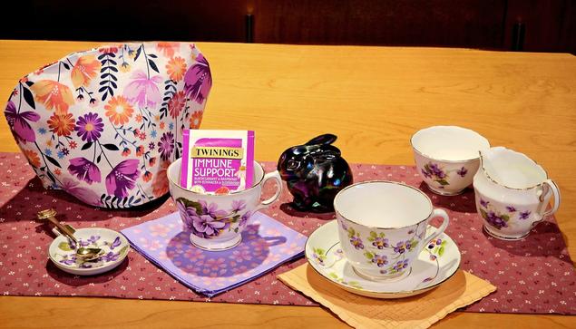 A tea tableau in shades of purple with orange accents, with china and linens decorated with violets, and a Fenton mercury glass bunny. The tea is a blackcurrant, raspberry, and elderberry herbal from Twinings.