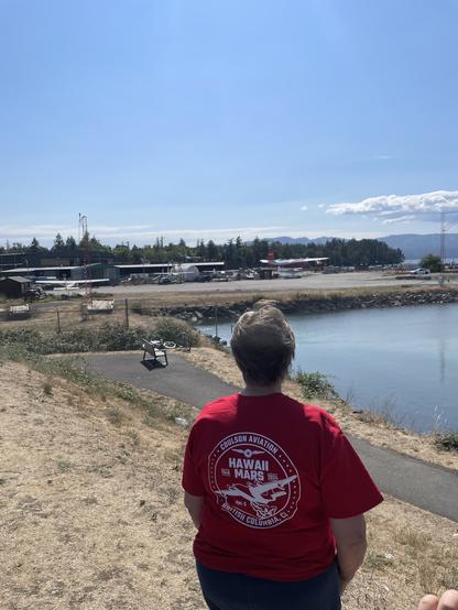 The Hawaii Mars is parked on a cement lot just above the water. A woman looks out toward it across a path and small bay. The woman is wearing a red Hawaii Mars commemorative TShirt
