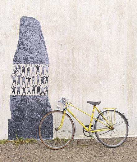 Menhir gris collé au mur : la moitié supérieure est portée par une pyramide humaine. À droite du menhir, un vélo jaune