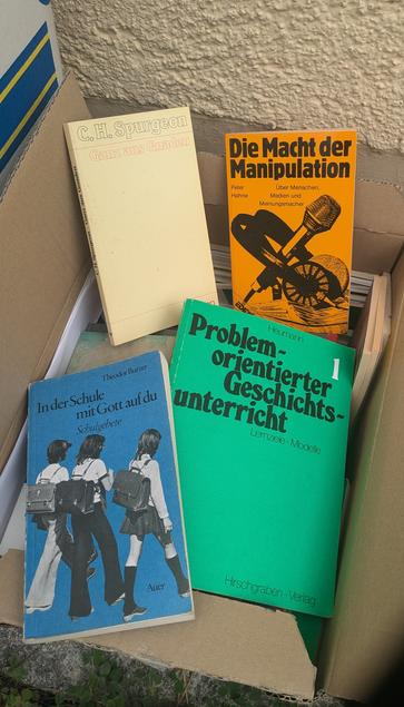 A cardboard box filled with books. The visible titles include "C.H. Spurgeon: Ganz aus Gnaden," "Die Macht der Manipulation" by Peter Hahne, and "In der Schule mit Gott auf du" by Theodor Bur