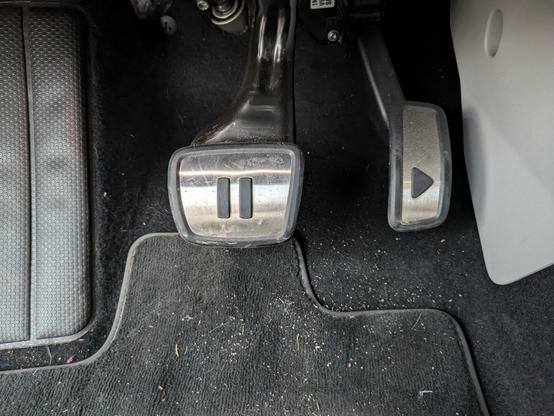 The brake and accelerator pedals in a VW electric campervan are shown. They are silver with black rubber markings. The accelerator has a right-facing arrow, so looks like a play button on a tape machine / music app UI. The brake has two vertical lines - the pause button.
