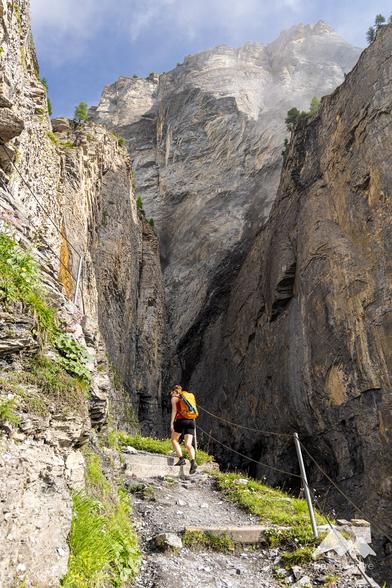 Woman wearing shorts, a red top and carrying an orange backpack, hiking up a mountain path in the Swiss Alps. Huge rock face in front.