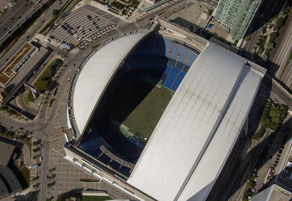 Aerial view of a large stadium with a partially closed retractable roof, surrounded by roads, buildings, and parking areas.