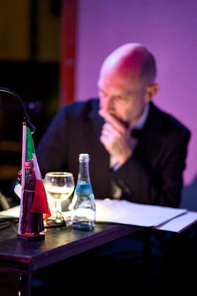 Im Hintergrund unscharf ein glatzköpfiger Mann im dunklen Anzug in Denkerpose. Auf dem Tisch vor ihm eine Plastikfigur des Schutzheiligen "San Pio", der eine italienische Fahne hält.