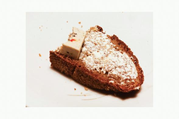 Photo of half a slice of wholemeal bread spread with margarine. The round side points to the lower right corner of the image. On the opposite site, there is a small chunk of tofu on it and there are bites, too. The sandwich is on a white plate, its edges are not visible, just some breadcrumbs and scratches around the sandwich.