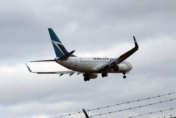 A blue and white WestJet plane takes off from Pearson International in Toronto. The plane is painted in the corporate colours: teal and blue on a white background. The sky is cloudy and grey. The top of a barbed wire fence is visible, bottom right. My photo, 2018.