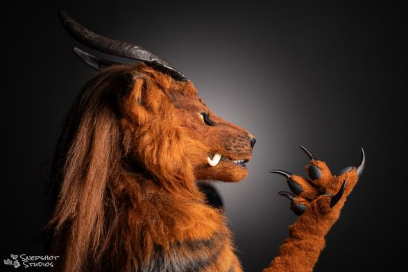 Side on bust portrait photo of a realistic foodog fursuit. Hand in front of the face as if giving a speech. A grey background with a circular gradient from light to dark.