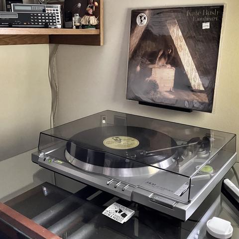 A turntable setup featuring Kate Bush's 'Lionheart' album. The turntable, an Akai model, is playing a record. Above it, the album cover for 'Lionheart' is displayed, showing a shadowy figure in a attic-like setting. On a shelf above, there's a calculator and a small hula dancer figurine.