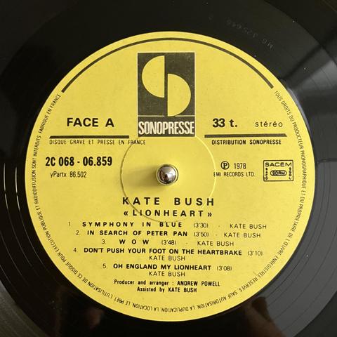 Close-up of a vinyl record label for Kate Bush's album 'Lionheart'. The label is yellow with black text, showing the Sonopresse logo and track listing for Side A. It includes songs like 'Symphony in Blue' and 'Oh England My Lionheart'. The label indicates it was produced in 1978 by EMI Records Ltd.