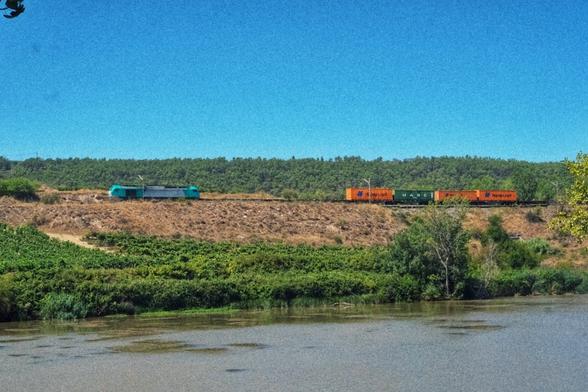 A container train pulled by an e-loco in turquoise livery, elevated above the water.