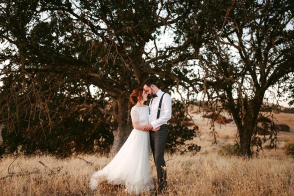 The image depicts a couple standing closely together in a dry, grassy field under the shade of a large, gnarled tree. The woman, dressed in a white lace wedding gown, leans into the man, who is wearing a white shirt, suspenders, and gray trousers. They are embracing tenderly, with the man's forehead resting gently on the woman's, creating an intimate and peaceful moment. The scene is set in a natural, somewhat rustic landscape, with the warm tones of the dried grass and the soft evening light adding to the romantic and timeless atmosphere. The background is softly out of focus, drawing attention to the couple as the central element of the image.