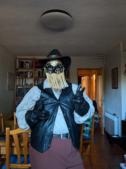 Gilead in costume as his LARP character Kennor Bruitascu, the cowboy bard, based on Orville Peck. I'm wearing a ren faire beige Shirt and brown trousers, a black leather vest, a black cowboy hat with a cow skull on the front, black leather gloves and an ornate facemask obscuring the upper half of my face, with golden fringe glued to the bottom rim obscuring the rest of my face. I'm trying my best to make hand pistols 🤣
