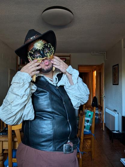Gilead in costume as his LARP character Kennor Bruitascu, the cowboy bard, based on Orville Peck. I'm wearing a ren faire beige Shirt and brown trousers, a black leather vest, a black cowboy hat with a cow skull on the front, black leather gloves and an ornate facemask obscuring the upper half of my face, with golden fringe glued to the bottom rim obscuring the rest of my face. I'm blowing a kiss towards he camera between the two halves of the fringe curtain of the mask, which I'm parting to the sides with my hands.