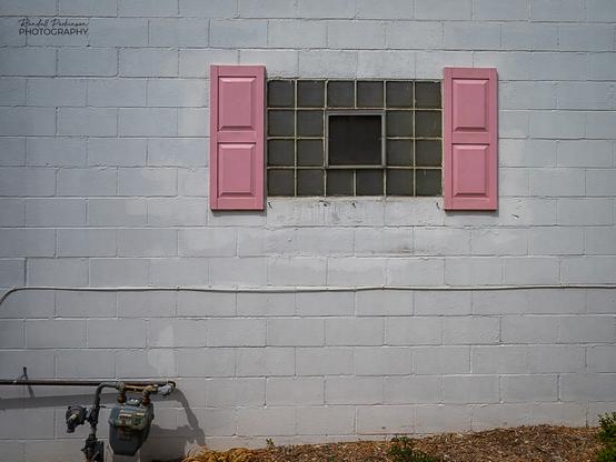 Pink shutters hang beside a glass block window in a white brick wall.