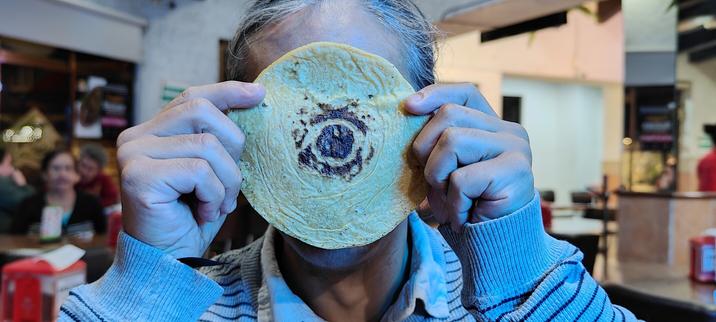 Un hombre sostiene delante de su cara una tortilla de maíz. En el centro de la tortilla se ve una mancha con la forma de un ojo.