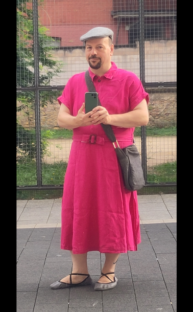 Selbstporträt in spiegelnder Scheibe. Mann mittleren Alters mit kräftiger Statur und dunklem Kinnbart steht in einer Fußgängerzone. Ich trage ein leuchtend pinkfarbenes Kleid aus Leinen. Oberteil geschnitten wie ein Kurzarm-Hemd mit Knöpfen, Rockteil wadenlang und ausgestellt. Dazu eine graue Tweed-Schiebermütze mit Herringbone-Muster, eine Umhängetasche im gleichen Muster, und graue Ballerinas (Gymnastikschuhe) mit Kreuzgummis.