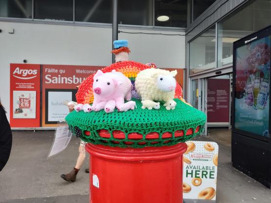 A photo of a postbox outside a Sainsbury's that has been yarn bombed with a crochet scene. On this side there is crochet grass and a pink pig with piglets and a sheep with little lambs. There is a crochet rainbow behind them.