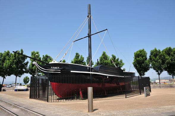 Replica of the paddle steamer "Comet" on display in Port Glasgow. The image shows a small paddle steamer viewed from its front left, so it is pointing left out of the frame. It has a red lower hull and black upper hull and mast and the name COMET is on the bow. It is surrounded by black railings and there is a line of small trees cutting across the frame in the background. The sky is blue.