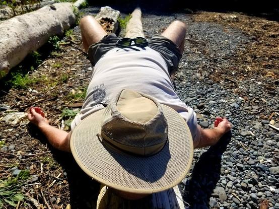 A man in white t-shirt & black shorts, laying with back flat on driftwood log, with khaki wide brim hat over his head/face. His arms are laying down on both sides of log. His legs are slightly bent at knee, planted feet on pebble beach on both sides of log. Black sunglasses are resting on his waist.