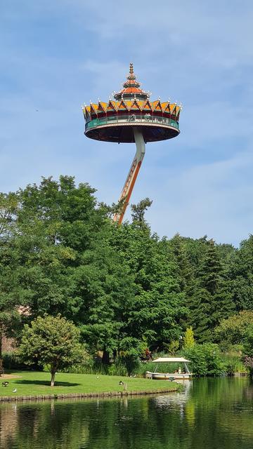 Theme park The Efteling's Pagode, a circular lookout platform in the style of an Asian pagoda on a hydraulic lifting arm, towering above a lake lined with trees, a small grass island and a white, roofed boat