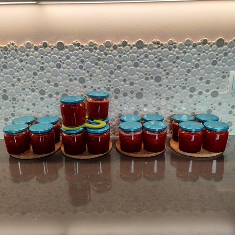 18 glass jars with blue lids, resting on round cork trivets, 4 to a trivet, except the second one that has 2 extra on top.

Jars are filled with strawberry jam.

Black, speckled counter. Bubble backsplash.