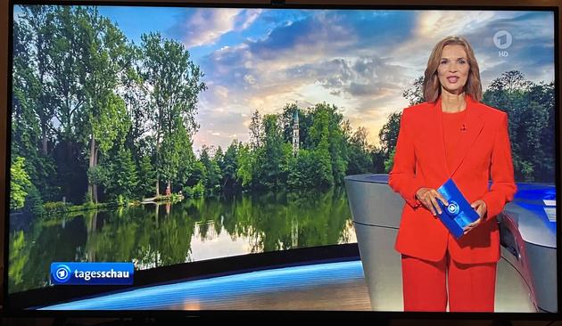 Ein Foto der Tagesschau auf einem Fernseher - zu sehen ist der See der Domaine Monrepos mit farbenfrohem bewölkten Himmel. Vor dem Foto steht die Moderatorin der Tagesschau und moderiert gerade das Wetter an