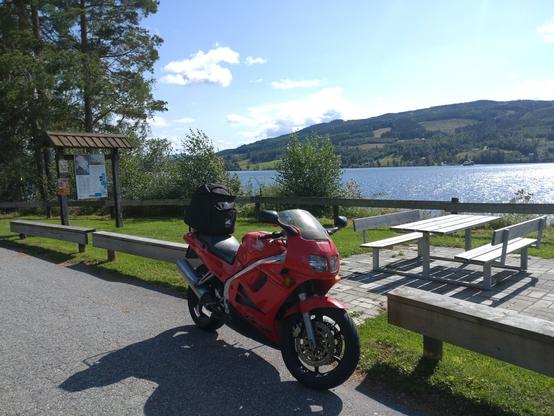 Rød motorsykkel på en rasteplass ved en sjø. I bakgrunnen er en ferge på vei over sjøen.