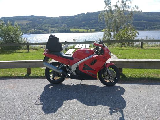 Rød motorsykkel på en rasteplass ved en sjø. I bakgrunnen er en ferge på vei over sjøen.
