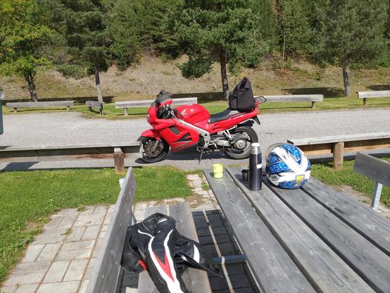 I forgrunnen en rasteplassbenk med termos og kopp og i bakgrunnen en rød motorsykkel.