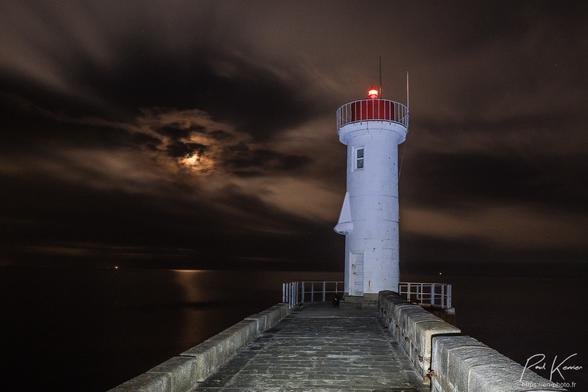 Photographie en couleur, de nuit, sous un ciel partiellement nuageux, laissant filtrer la lune on peut y voir au bout d'une jetée en maçonnerie de pierres, un petit phare avec un feu rouge au sommet. un nez a été ajouté sur la tour du phare, alors que la porte d'entrée représenterait la bouche et une fenêtre au-dessus du nez, un œil. L'ensemble me fait penser. à un moaï pasquouan.