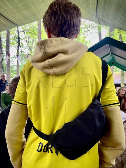 das foto im hochformat zeigt einen jugendlichen von hinten, konkret das fußballtrikot, das er anhat.
es ist ein gelbes trikot und unterhalb seiner umgehängten schwarzen schultertasche lässt sich das "dort" von "dortmund" lesen.
als fan des fußballspielers marco reus hat der jugendliche mit einem schwarzen marker den namen des spielers reus in großbuchstaben über die schulterfläche geschrieben, darunter mittig die ziffer und spielernummer 11.
im hintergrund sind weitere leute in einem waldgebiet zu erkennen.