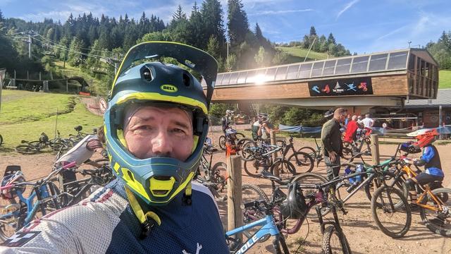 Photo of me with a full-face helmet in front of a lot of mountain bikes and a lift. Blue sky, a mountain behind me, tall trees. Two people on the right, probably father and son.
