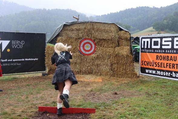 A woman in a dark outfit and plaid skirt is performing a throw toward a target marked on a haystack. The backdrop includes banners for local businesses. The setting appears to be an outdoor event in a rural area with a misty landscape.