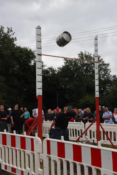 A crowd watches as a competitor attempts to throw a keg over a high bar during a strength competition. The keg is in mid-air, and participants are dressed in athletic attire, with some wearing kilts. A cloudy sky is visible in the background.