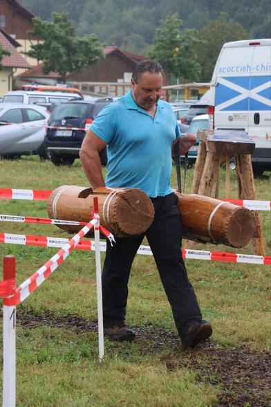 A disabled man is carrying two large wooden logs in a grassy outdoor area, with a red and white striped barrier in the foreground. It appears to be raining, and there are parked cars and buildings in the background.