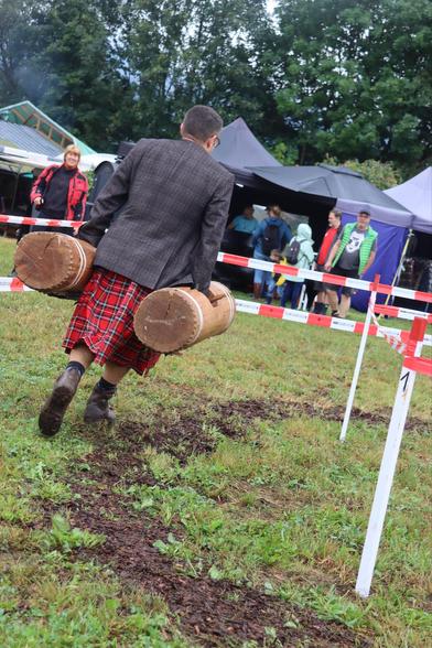 A person in a kilt is carrying two heavy wooden barrels while walking on a grassy field. In the background, there are tents and spectators watching the event.