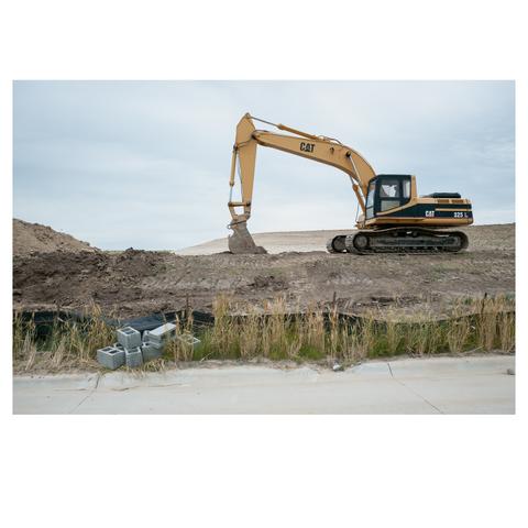 A backhoe sits on a bulldozed hill above a new road. A pile of cinder blocks sits on the side of the hill in the left part of the frame. A larger hill and pale blue sky lie behind the backhoe.