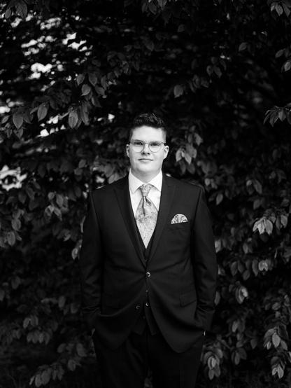This image is a black-and-white portrait of a groom standing in front of a dense backdrop of leafy trees or bushes. The groom is dressed in a formal suit, including a tie and a pocket square, which adds a touch of elegance to his outfit. He has short, neatly styled hair and is wearing round glasses, giving him a refined and thoughtful appearance. His hands are casually placed in his pants pockets, and he is looking directly at the camera with a calm and composed expression. The blurred background emphasizes the groom, making him the clear focal point of the image. The overall mood is classic and timeless, capturing a quiet moment of confidence and reflection before the wedding ceremony.