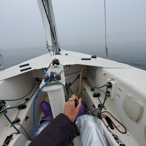 View of steering & sails controls from my pilot/captain seat. My left hand is on steering stick & I've pulled it to the right. In front of boat is foggy ocean.