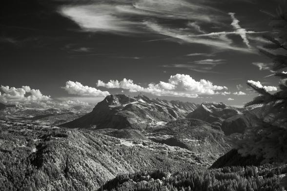 Black and white, infrared photography: A valley and mountains flow from left to right through the image, clouds are dotting the dark grey sky. A fir tree’s branches out of focus frame the image to the right.