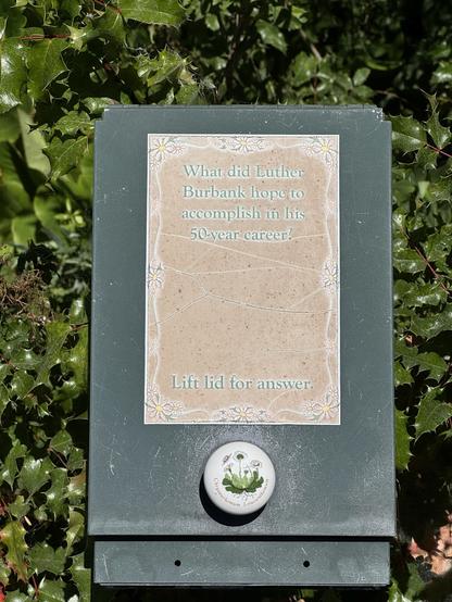 A sign asking, "What did Luther Burbank hope to accomplish in his 50-year career?" with instructions to "Lift lid for answer," placed on a green backdrop.