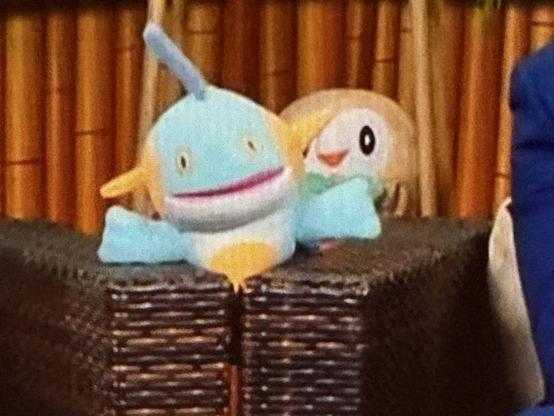 A plush toy of a light blue aquatic character with orange fins and a smile is placed next to a green and white roundish bird plush toy, set against a backdrop of bamboo.