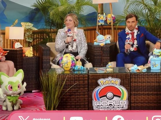 A Pokémon-themed stage at an event features two hosts seated on a wicker couch. The first host, dressed in a sparkling jacket, holds a microphone and sits next to a colorful Pokémon ball. The second host, in a blue suit, looks serious.