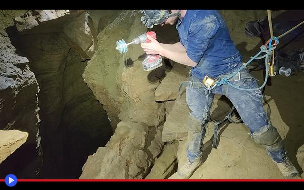 Speleologo si sporge sopra un baratro oscuro in un ambiente sotterraneo, tenendo in mano un trapano con un rocchetto di filo all'estremità.