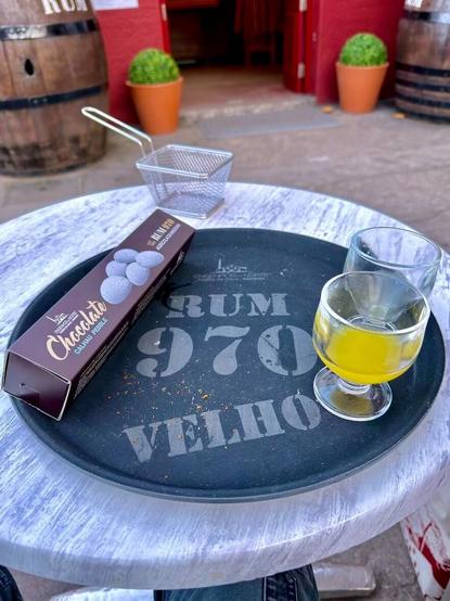 Das Bild zeigt ein rundes, schwarzes Tablett mit der Aufschrift „Rum 970 velho“ und zwei kleinen Gläsern mit dem Rest eines gelblichen Getränks, einem Poncha. Daneben liegt eine längliche Packung mit aufgedruckten Schoko-Mandeln.