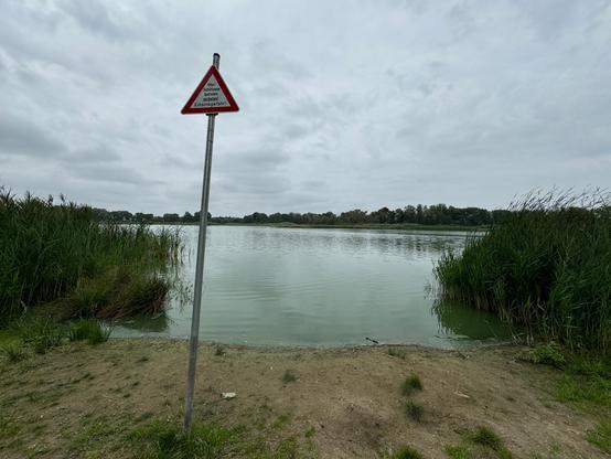 Die Badestelle am (Löhmer) Haussee in Werneuchen/Seefeld, an einem kühlen Sommertag, unter bewölktem Himmel. Am Ufer hängt an einem schiefen Ständer ein dreieckiges, rot umrandetes, Schild mit der Aufschrift: "Ufer/Schilfzone betreten verboten! Einsinkgefahr!"