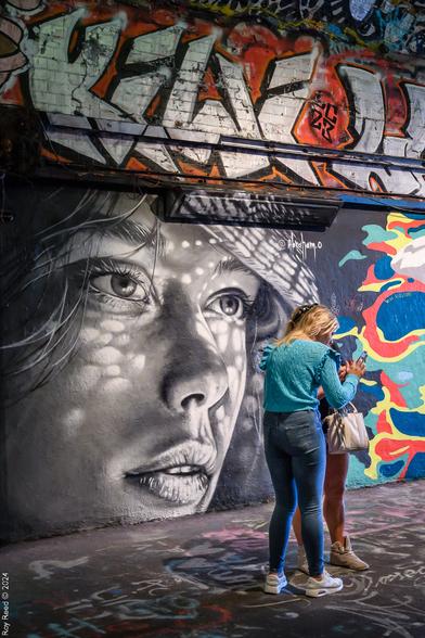 Two young women reviewing a selfie while standing in front of a street art portrait of a girl's face painted in black and white.